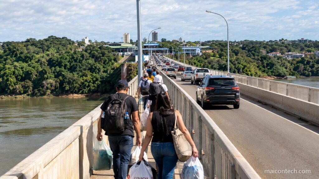 Pessoas a atravessar a pe uma ponte internacional de fronteira carregando sacos de compras, com transito de carros ao lado e o rio por baixo
