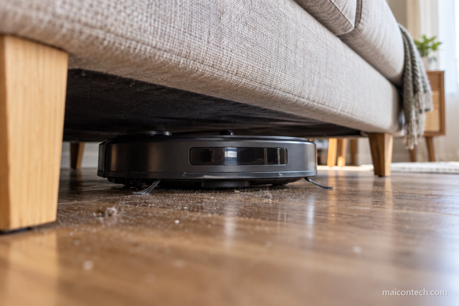 Robo aspirador visto ao nivel do chao a entrar por baixo de um sofa, com po e cotao visiveis no soalho